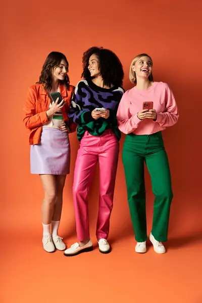 Three young, beautiful women in stylish attire from different cultures stand united on an orange background. — Stock Photo