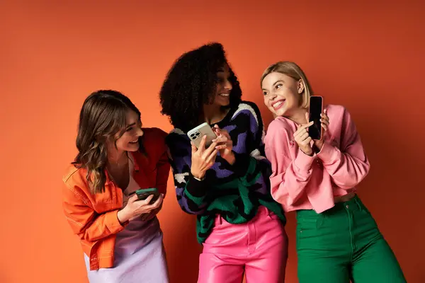 Three young, beautiful, multicultural women in stylish vibrant clothes standing together, looking at their cell phones. — Stock Photo