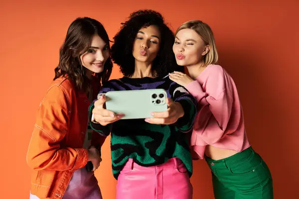 Three young women in stylish attire stand together, holding a cell phone, capturing a moment of friendship and cultural diversity. — Stock Photo