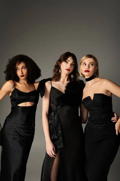 A diverse trio of young women in elegant black dresses posing gracefully for a stylish portrait on a grey background. — Stock Photo