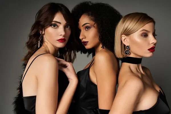 Three stunning women, of diverse ethnicities, striking poses in elegant black dresses against a grey backdrop. — Stock Photo