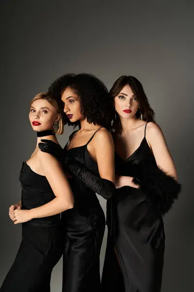 Три молодих, красивих міжрасових жінки стоять єдиними, демонструючи різноманітність в елегантних чорних сукнях, на сірому фоні. — стокове фото