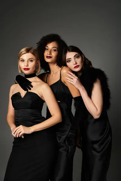 Three young, beautiful interracial women in elegant black dresses posing gracefully against a grey background. — Stock Photo
