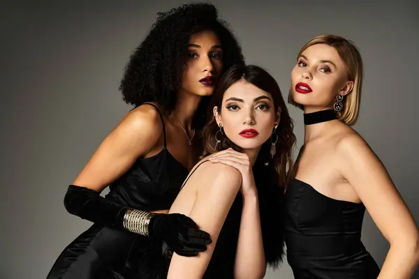 Three beautiful, diverse women in elegant black dresses posing gracefully for a fashion shoot on a grey background. — Stock Photo