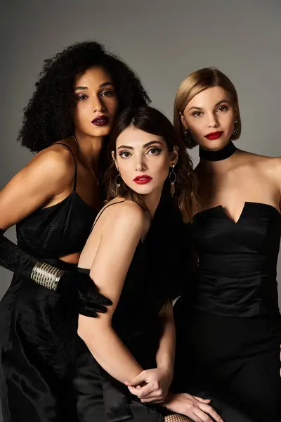 Три молодих красивих жінок різних національностей витончено позують у чорних сукнях на сірому тлі. — стокове фото