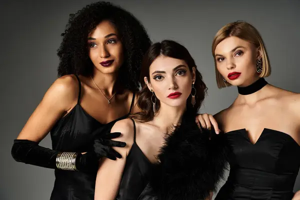 Three young, beautiful women of different ethnicities posing gracefully in elegant black dresses against a grey backdrop. — Stock Photo