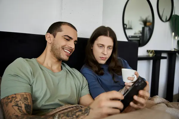 A young gay couple relaxes in bed, enjoying coffee and each others company. — Stock Photo