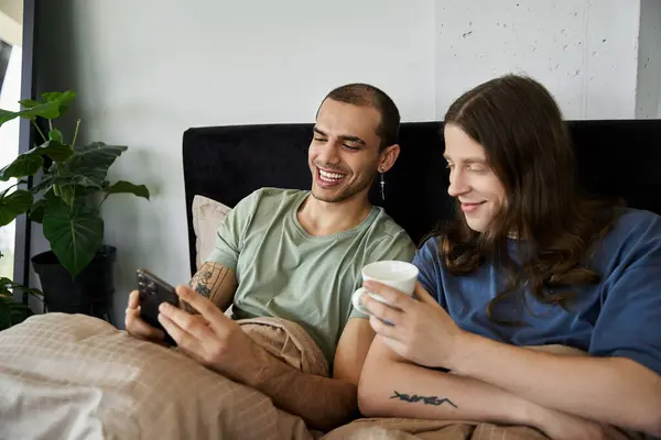 A young gay couple smiles and laughs together in bed, enjoying their morning coffee and looking at their phone. — Stock Photo