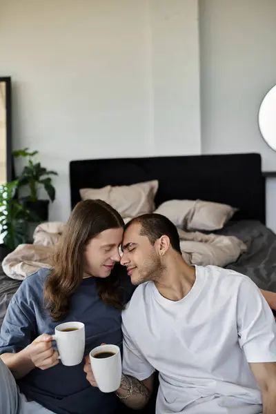 Um jovem casal gay se senta em uma cama, desfrutando de café juntos em um ambiente acolhedor quarto. — Fotografia de Stock