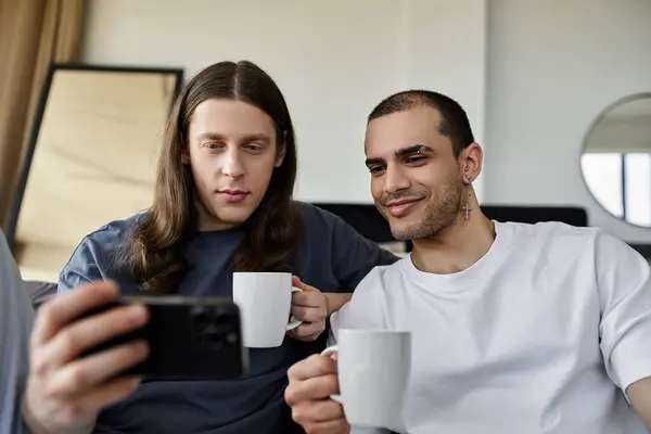 Una giovane coppia gay gode di una mattina accogliente insieme nella loro camera da letto, condividendo un drink caldo e guardando il loro telefono. — Foto stock