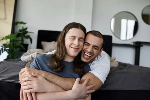 A young gay couple smiles and laughs as they embrace each other on a bed in a bedroom. — Stock Photo