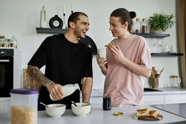 Dos hombres gay disfrutan del desayuno juntos en su cocina moderna; uno vierte leche en un tazón mientras que el otro sonríe con tostadas. - foto de stock