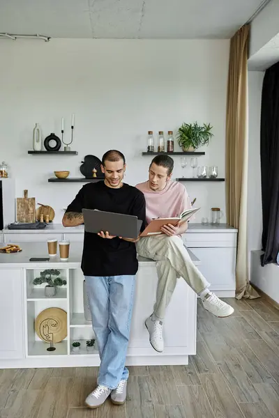 A young gay couple spends time together in a modern kitchen, one working on a laptop while the other reads a book. — Stock Photo