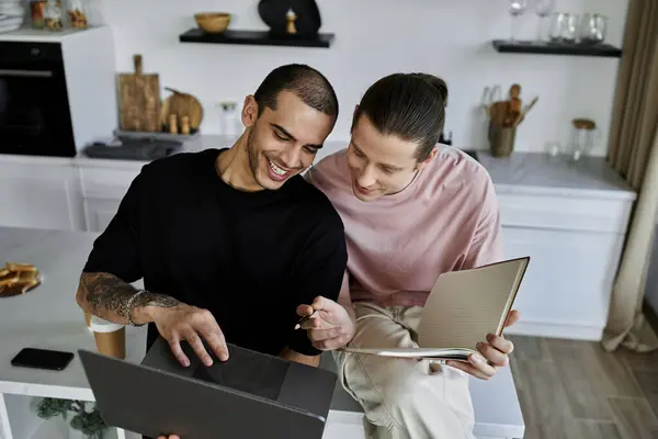 Two young men are sitting in a modern kitchen, using a laptop and a notebook together. They seem to be enjoying each others company. — Stock Photo