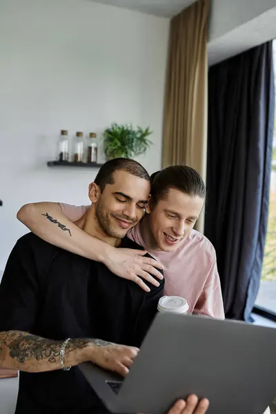 A young gay couple cuddles together as they browse the internet on a laptop in their modern home. — Stock Photo