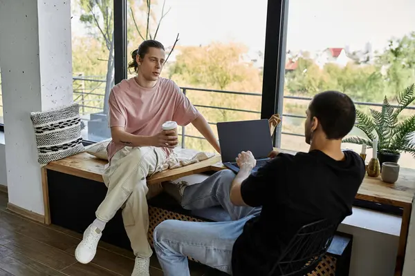 Two young men relax on a window seat in a modern home, enjoying coffee and conversation. — Stock Photo