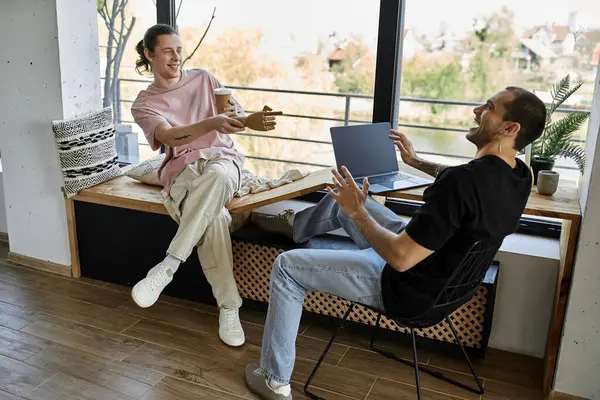 Um jovem casal gay desfruta de uma pausa para o café em sua casa moderna, rindo e conversando enquanto se usa um laptop. — Fotografia de Stock