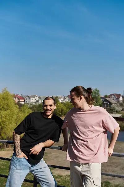 Dos jóvenes, vestidos casualmente, se paran uno al lado del otro en una barandilla con vistas a un barrio suburbano en un día soleado. - foto de stock