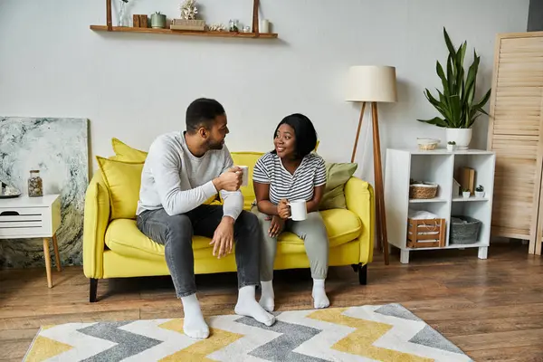 A loving African American couple sits on a yellow couch, enjoying coffee together in their home. — Stock Photo