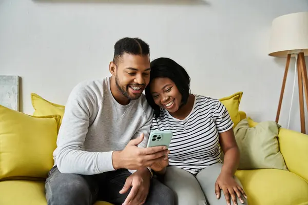 A couple sits on a yellow couch, laughing while looking at a phone. — Stock Photo