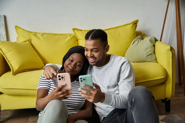 A loving couple sits on a yellow couch looking at their phones. — Stock Photo