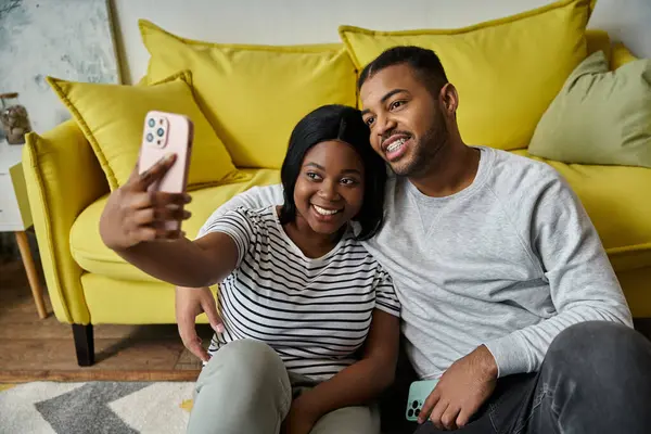 A loving African American couple takes a selfie on their yellow sofa at home. — Stockfoto