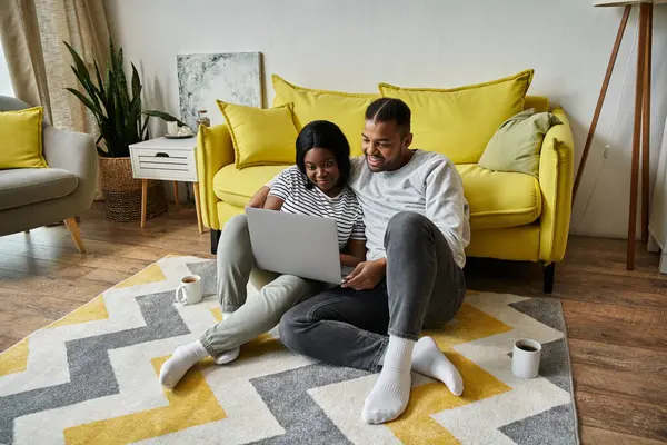 An African American couple sits on a rug in their living room, looking at a laptop together. — Stock Photo