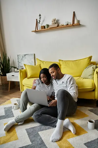 Una pareja afroamericana se sienta en una alfombra frente a un sofá amarillo, usando un ordenador portátil juntos. - foto de stock