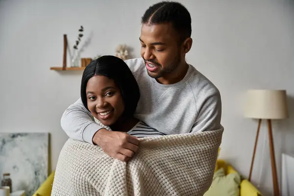 A loving couple cuddles under a blanket in their home, with the man holding his partner close. — Stock Photo