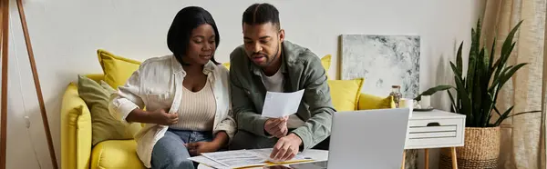 An African American couple sits on a yellow sofa, reviewing paperwork and using a laptop. — Stock Photo