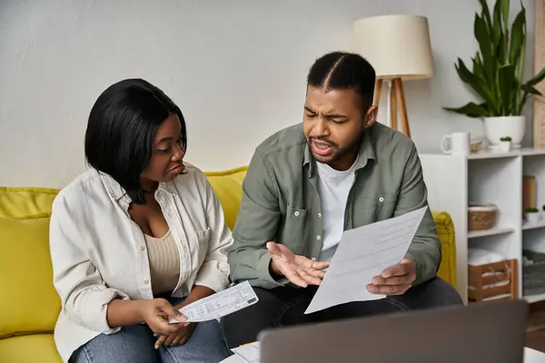 Un couple afro-américain est assis sur un canapé à la maison, examinant la paperasserie et discutant des finances. — Photo de stock