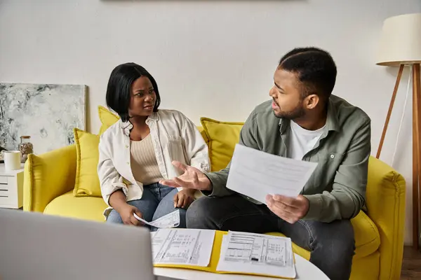 Una coppia afroamericana amorevole discute le loro finanze mentre si siede su un divano giallo a casa. — Foto stock