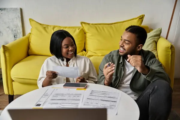 Una coppia afroamericana amorevole si siede su un divano giallo a casa, ridendo durante la revisione dei documenti. — Foto stock