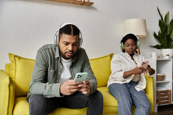 Una coppia afroamericana si rilassa su un divano giallo a casa, ciascuno assorto nei loro telefoni. — Foto stock