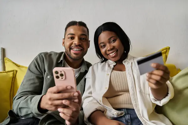 A loving African American couple uses a phone and credit card to shop online at home. — Stock Photo
