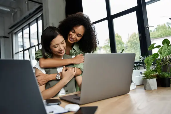 Two women, a lesbian couple, embrace while working on a laptop in a bright office. — Stock Photo
