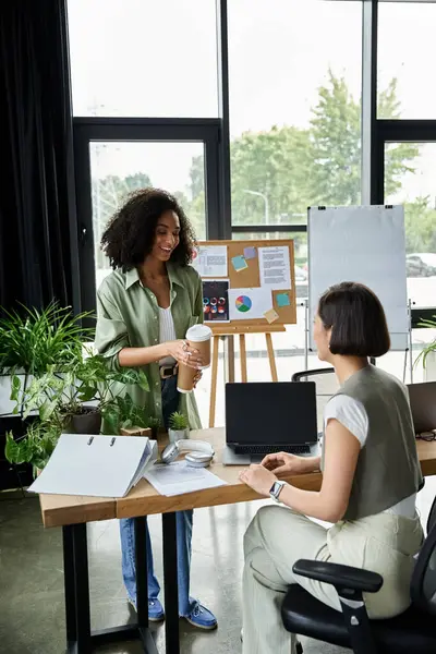 Lesbian couple working in modern office; one standing with coffee, one at desk on laptop. — Stock Photo