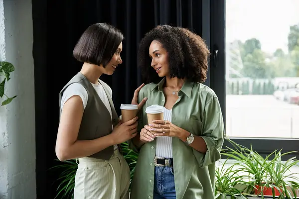Две женщины в офисе, держат чашки кофе и разговаривают. — стоковое фото