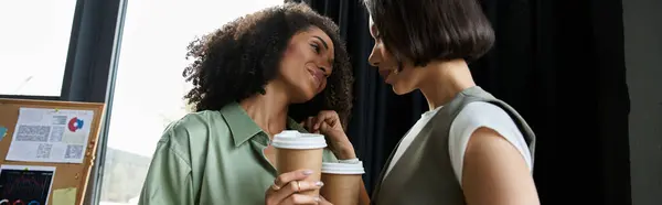 Zwei Frauen halten Kaffeetassen in der Hand und schauen einander in einem modernen Büro liebevoll an. — Stockfoto