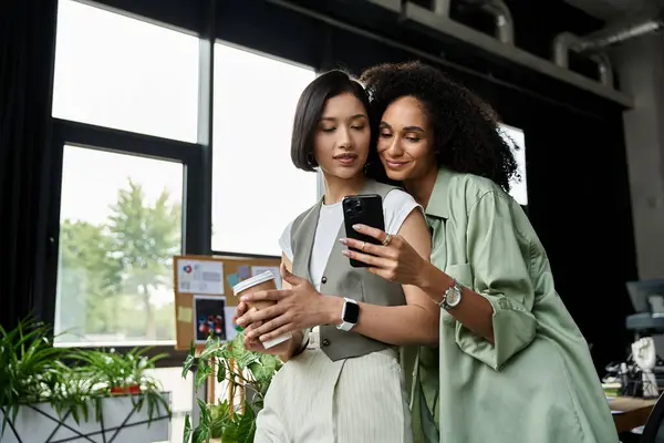 Two women working in an office, standing close and looking at a phone, enjoying each others company. — Stock Photo