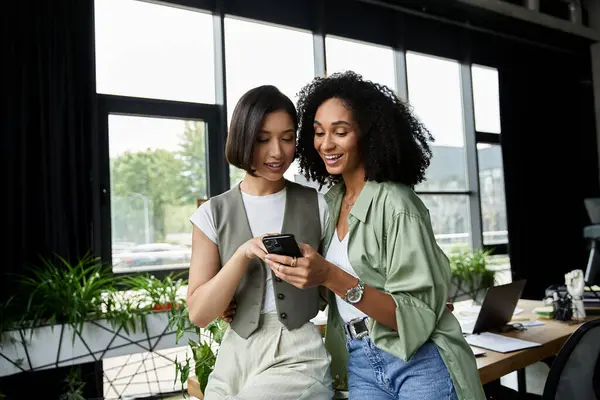 Two women in an office look at a smartphone together. — Stock Photo