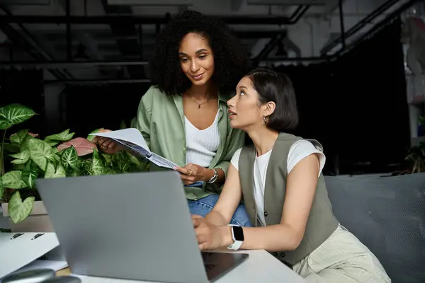 Two women, dressed in green, work on a project together at their office desk. — Stock Photo
