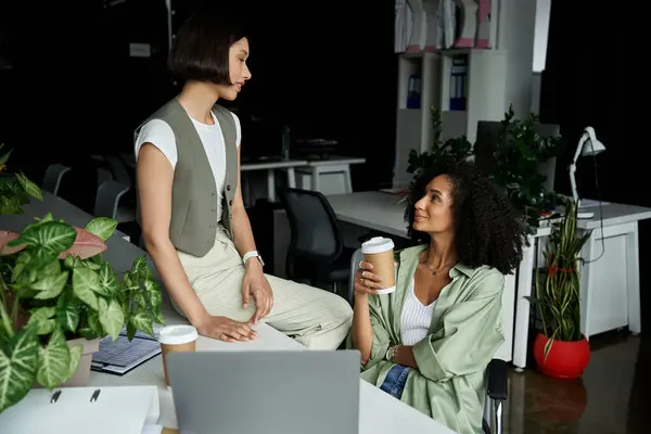 Two women, in an office setting, share a cup of coffee and work together. — Stock Photo