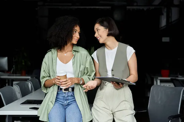 Two women in office, one with a clipboard, the other with coffee, talking. — Stock Photo