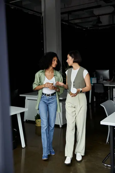 Zwei Frauen gehen im Büro spazieren und unterhalten sich, während sie Kaffeetassen halten. — Stockfoto