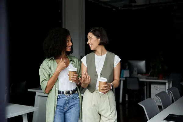 Two women in an office setting, holding coffee cups and smiling at each other. — Stock Photo