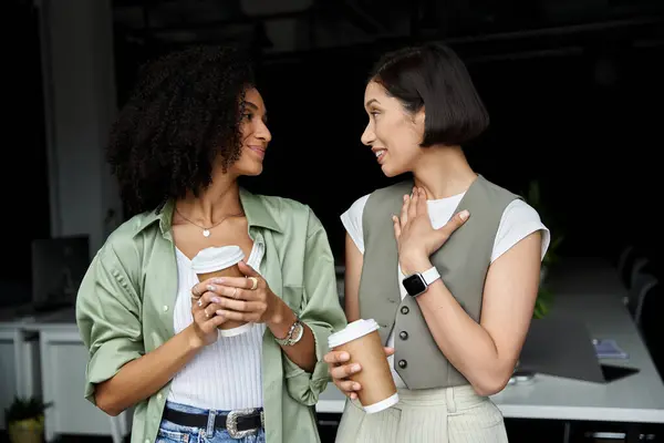 Two women stand in an office setting, holding coffee cups and talking to each other. — Stock Photo