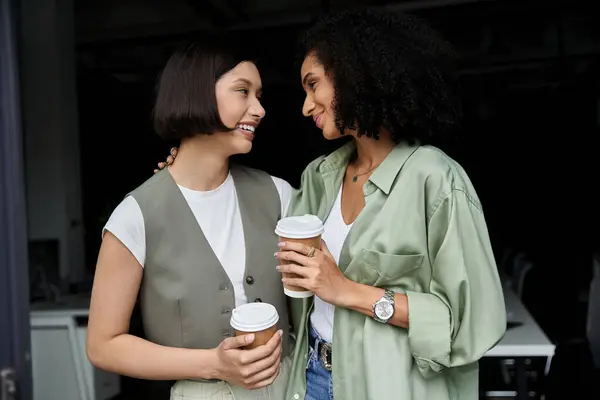 Duas mulheres, um casal lésbico, se reúnem em um escritório, sorrindo e segurando xícaras de café. — Fotografia de Stock