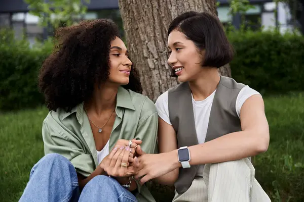 Two women are sitting together under a tree, holding hands and smiling. — Stock Photo