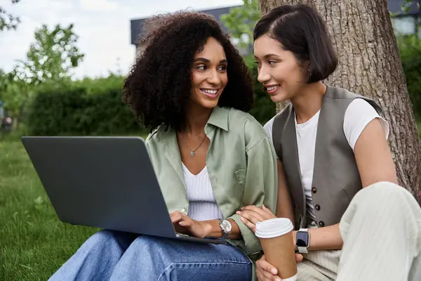 Two women work on a laptop outdoors, smiling and leaning in towards each other. — Stock Photo